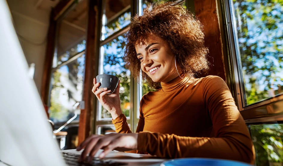 Lächelnde junge Frau sitzt mit einer Tasse Kaffee vor ihrem Laptop.